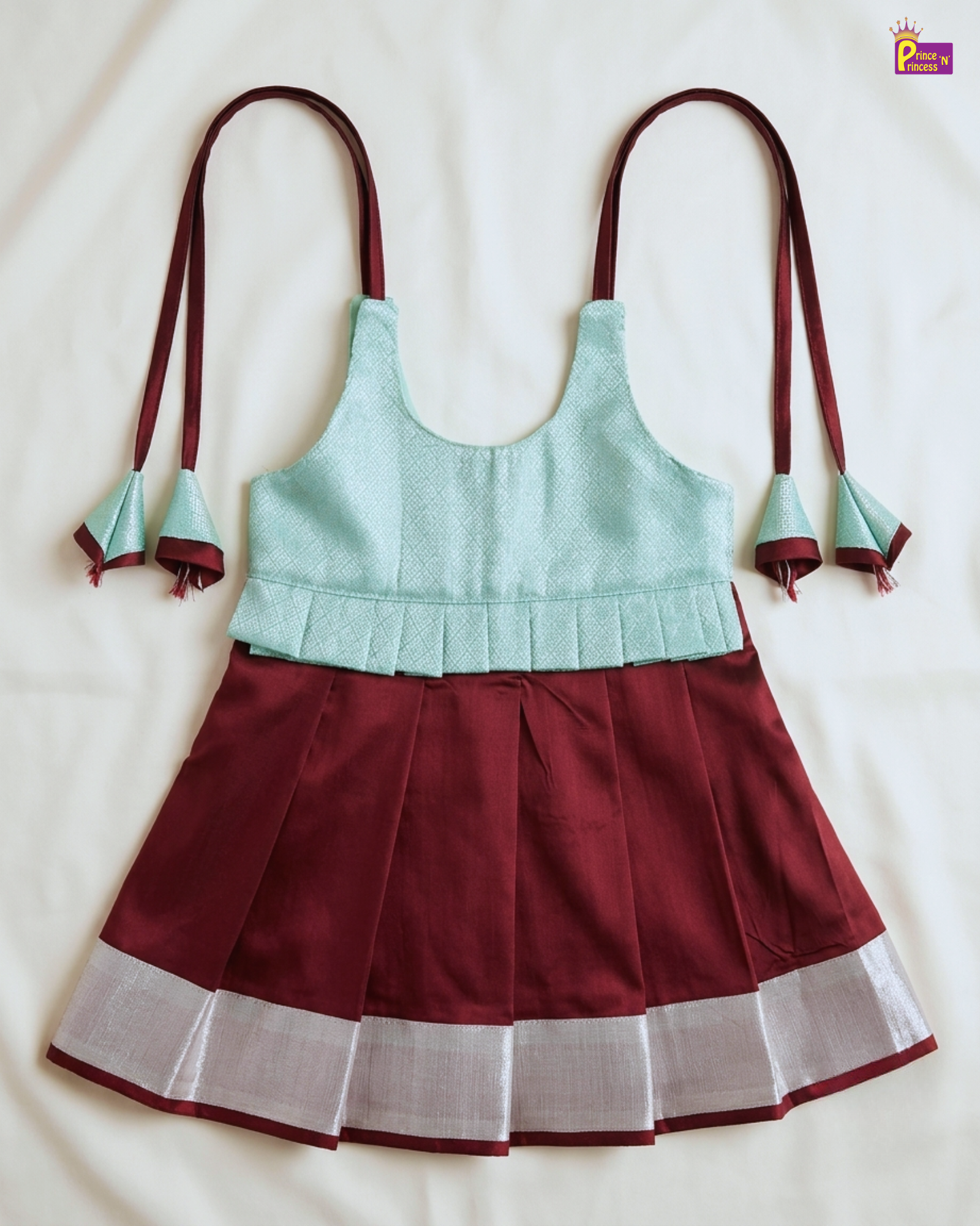 A traditional Indian salwar suit set featuring a pleated maroon skirt with a silver zari border, a light mint green sleeveless top, and matching maroon ties with silver decorative tassels, laid flat on a white fabric background.