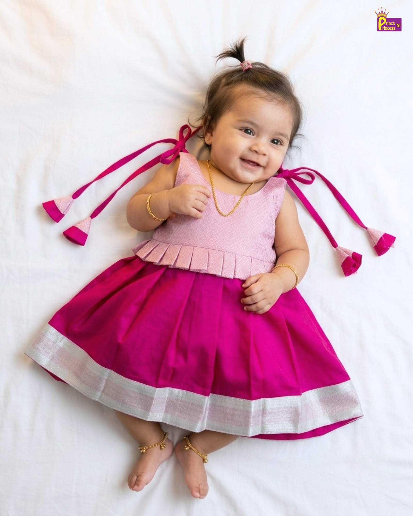 A baby girl lying on a white sheet, wearing a pink sleeveless dress with a pleated bodice, a bright magenta skirt with a silver border, and matching pink hair ties with tassels.