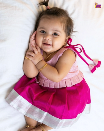 A baby girl lying on a white bed wearing a bright pink traditional dress with a pleated light pink top, a darker pink skirt, and a silver border.
