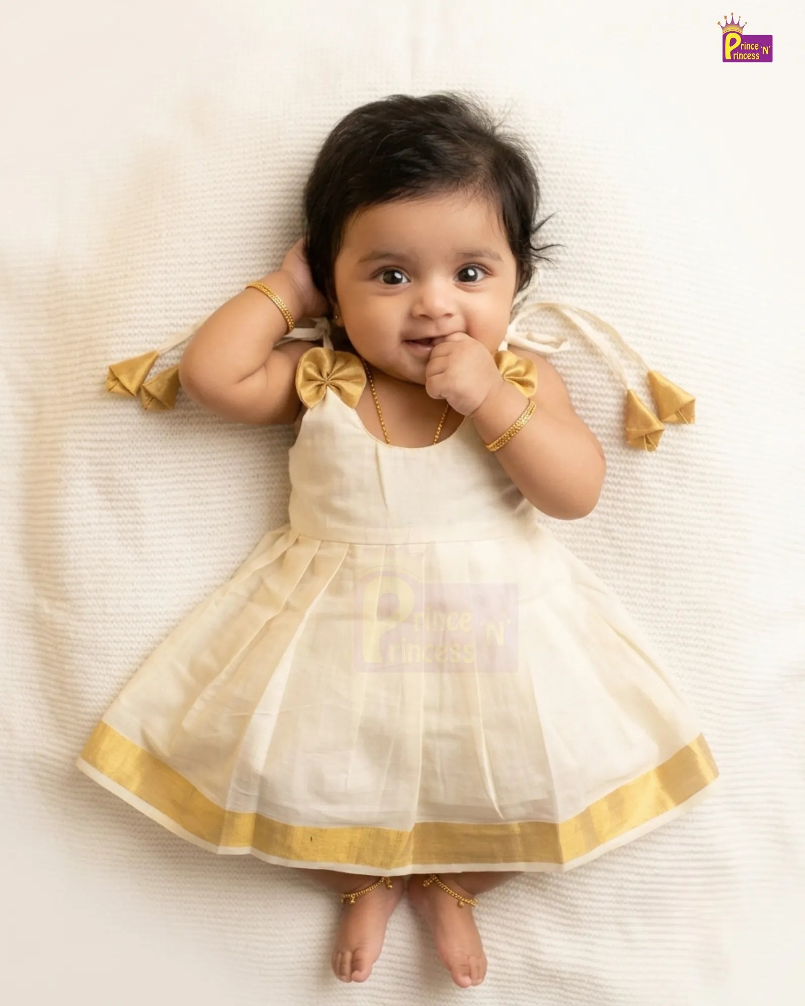 A baby girl lying on a white textured blanket, wearing a sleeveless cream-colored traditional dress with gold trim and bow details on the shoulders.