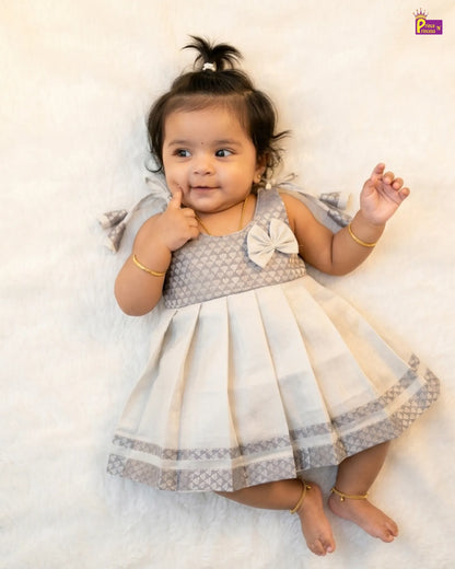 Baby in a gray and white dress with a bow, lying on a white surface.