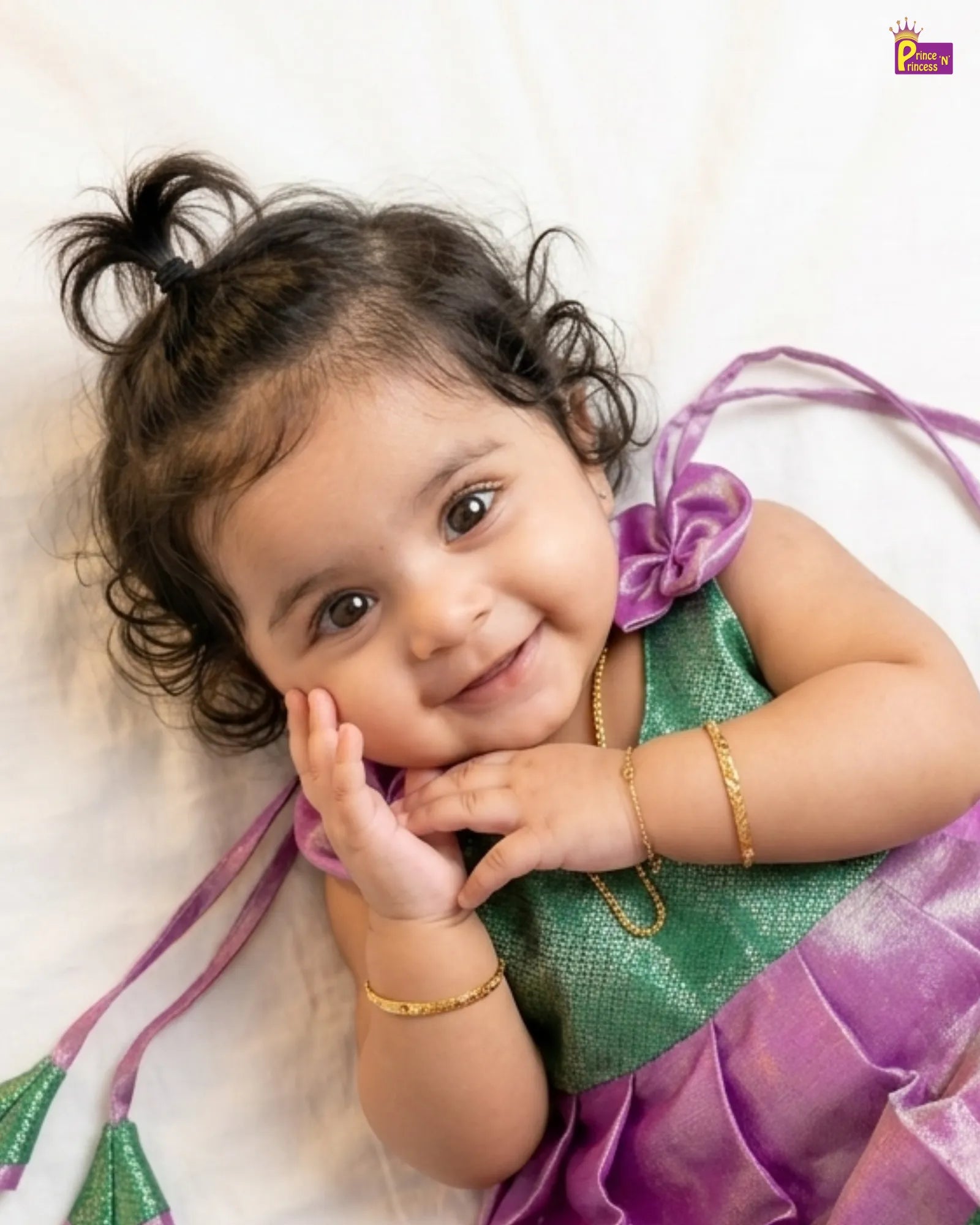 Child wearing a green and purple traditional outfit with a gold bracelet, lying on a white background.
