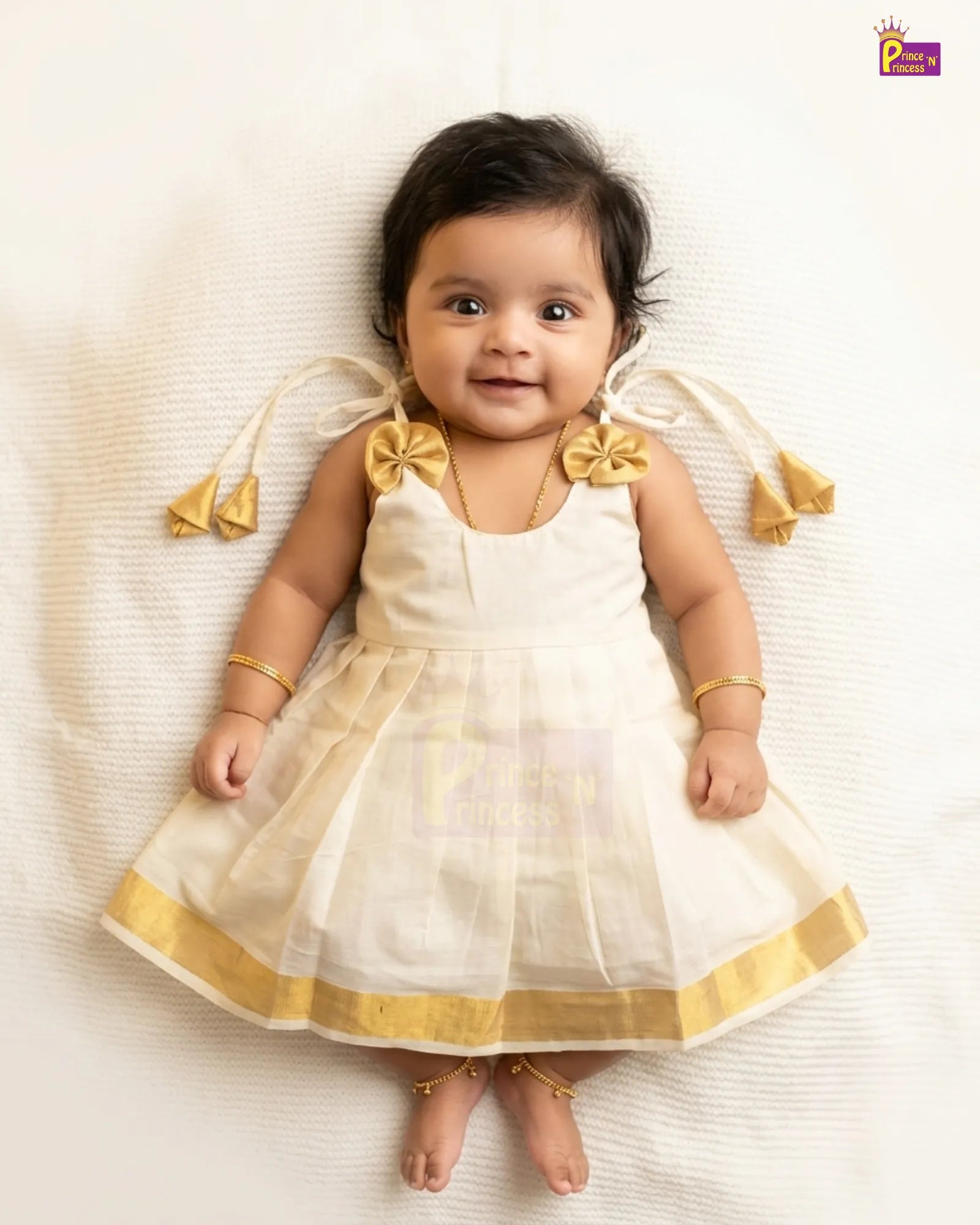 A baby girl lying on a white textured blanket, wearing a sleeveless white dress with gold trim, matching gold bow accessories on her shoulders, and gold bracelets and anklets.