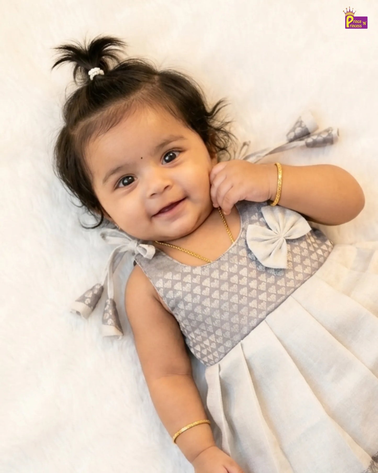Baby in a gray and white dress with a bow, lying on a white surface.