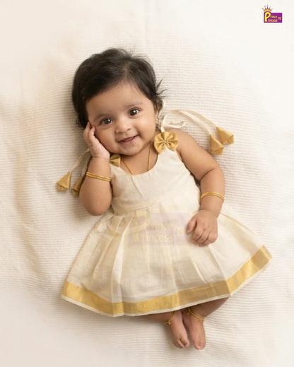 A baby girl lying on a textured cream blanket, wearing a sleeveless cream and gold dress with a bow on the shoulder and gold bangles.