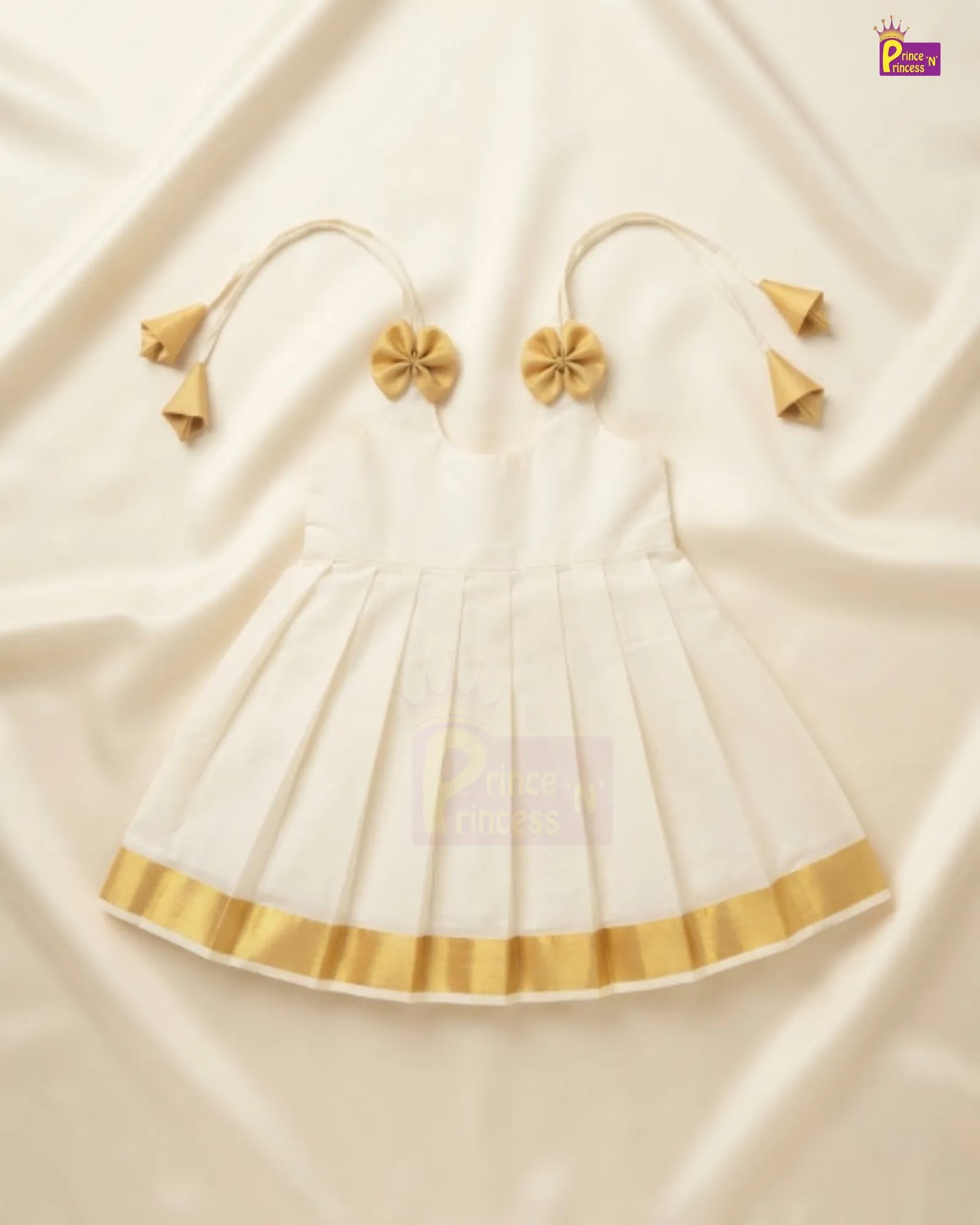A white sleeveless baby dress with gold trim and matching gold tie-shoulder accessories, laid flat on a cream-colored satin fabric background.