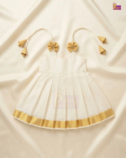 A white sleeveless baby dress with gold trim and matching gold tie-shoulder accessories, laid flat on a cream-colored satin fabric background.