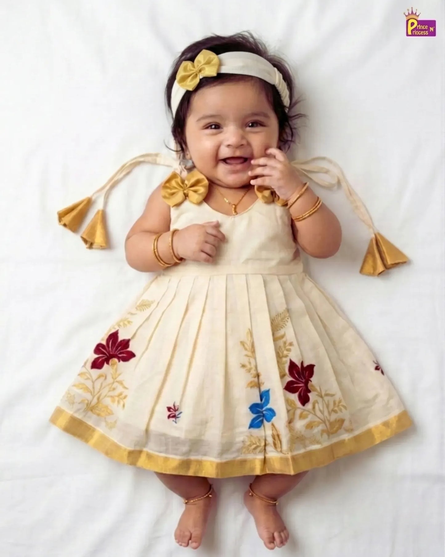 A baby girl lying on a white sheet wearing a cream-colored sleeveless dress with gold floral embroidery, a matching headband with a yellow bow, and gold jewelry.