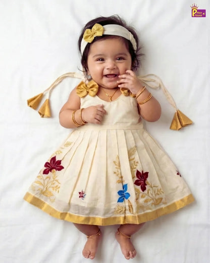 A baby girl lying on a white sheet wearing a cream-colored sleeveless dress with gold floral embroidery, a matching headband with a yellow bow, and gold jewelry.