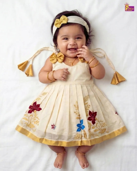 A baby girl lying on a white sheet wearing a cream-colored sleeveless dress with gold floral embroidery, a matching headband with a yellow bow, and gold jewelry.