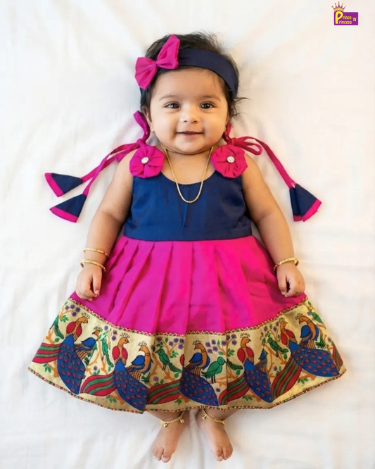 A toddler girl lying on a white sheet wearing a two-tone navy blue and hot pink dress with a peacock patterned hem, a matching pink bow headband, and gold jewelry.