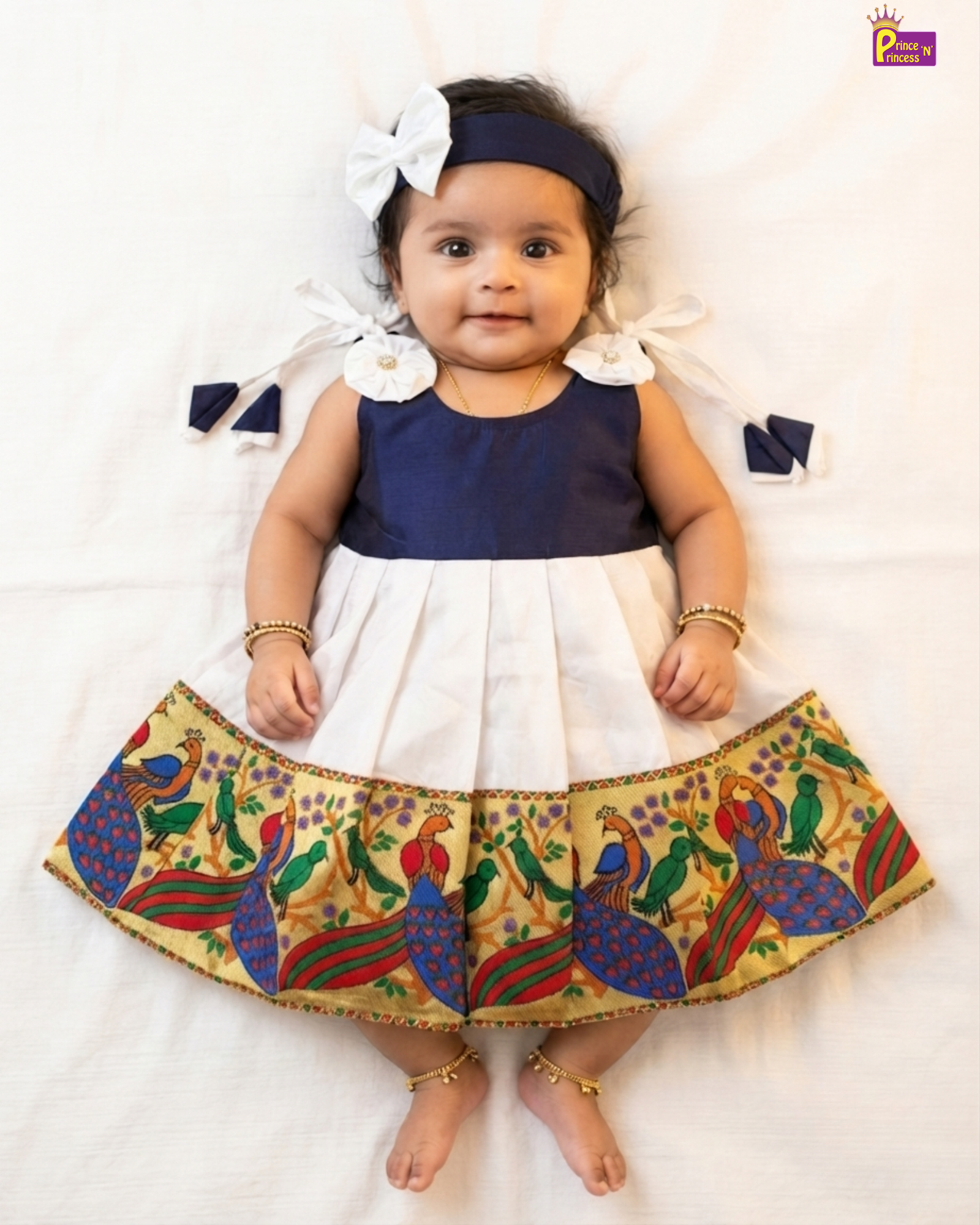 A baby girl lying flat on a white sheet wearing a navy blue and white two-tone dress with a colorful peacock patterned skirt, a navy headband with a white bow, and gold bangles and ankle bracelets.