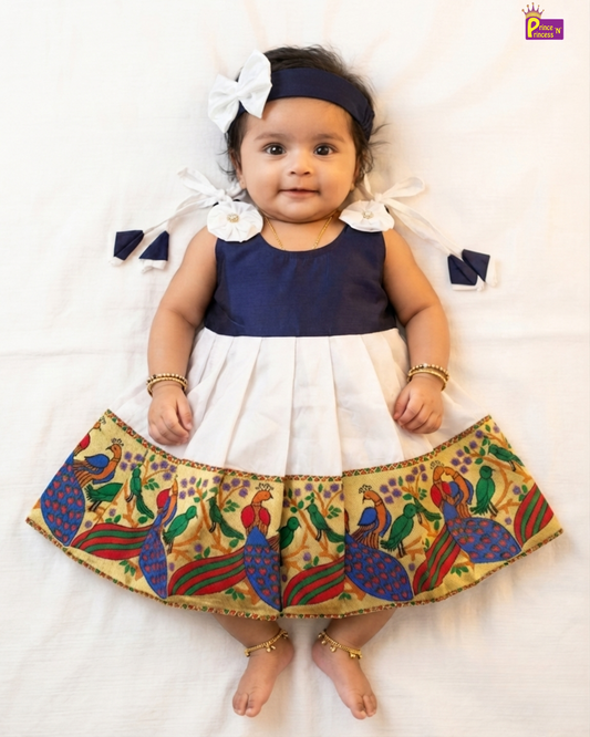 A baby girl lying flat on a white sheet wearing a navy blue and white two-tone dress with a colorful peacock patterned skirt, a navy headband with a white bow, and gold bangles and ankle bracelets.