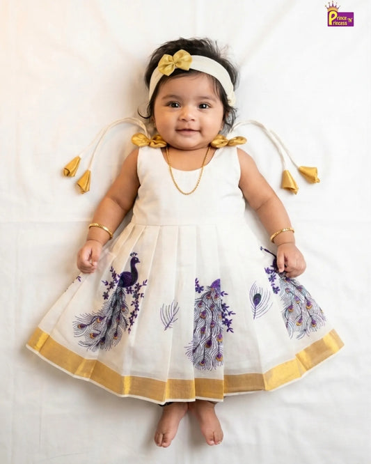 A baby girl wearing a white sleeveless dress with purple peacock prints and gold trim, a matching white headband with a gold bow, and gold jewelry, lying on a white fabric background.
