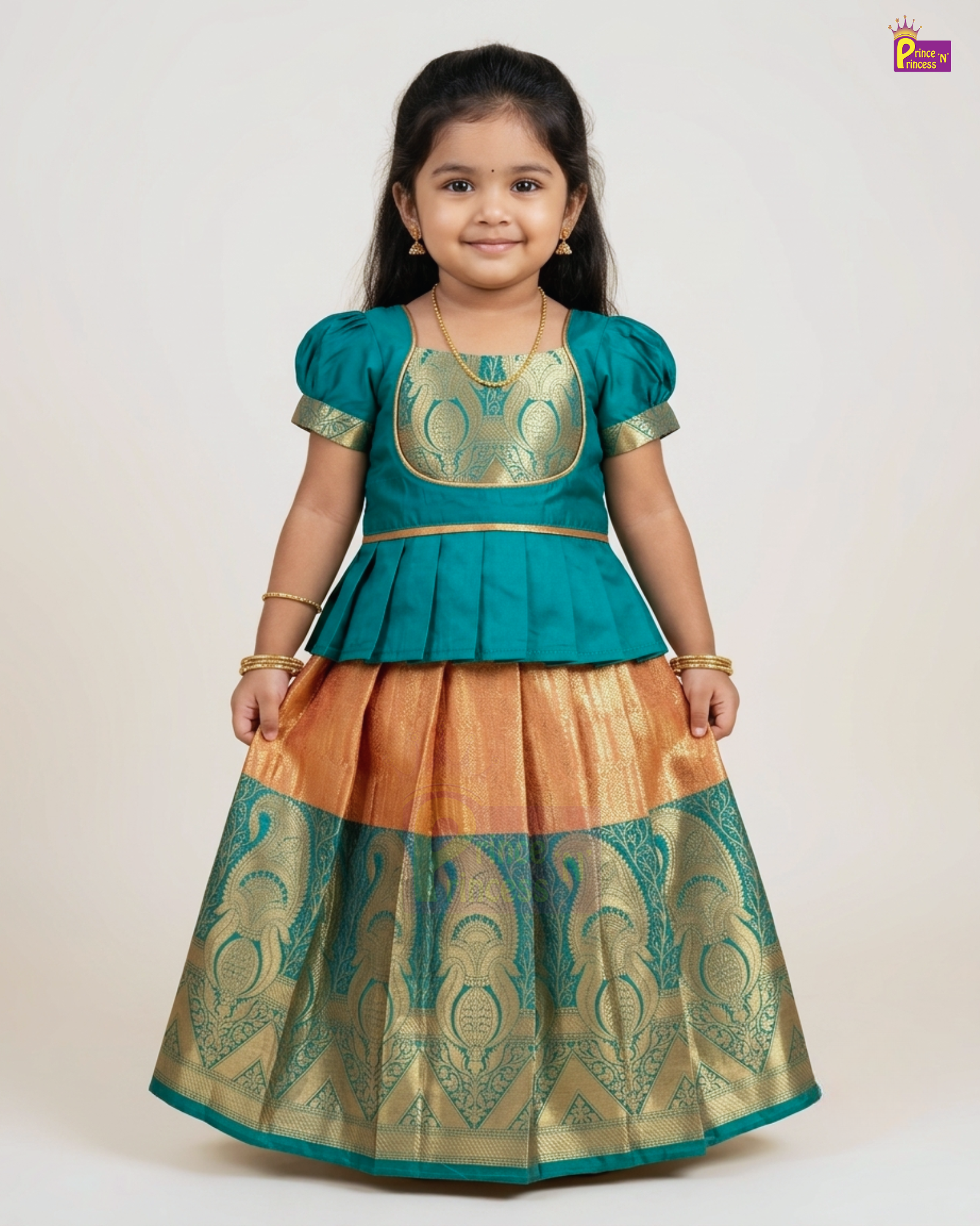A young girl modeling a traditional Indian ethnic outfit consisting of a teal blue peplum top with gold embroidery and a matching flared skirt in gold and teal with woven patterns, set against a plain light background.