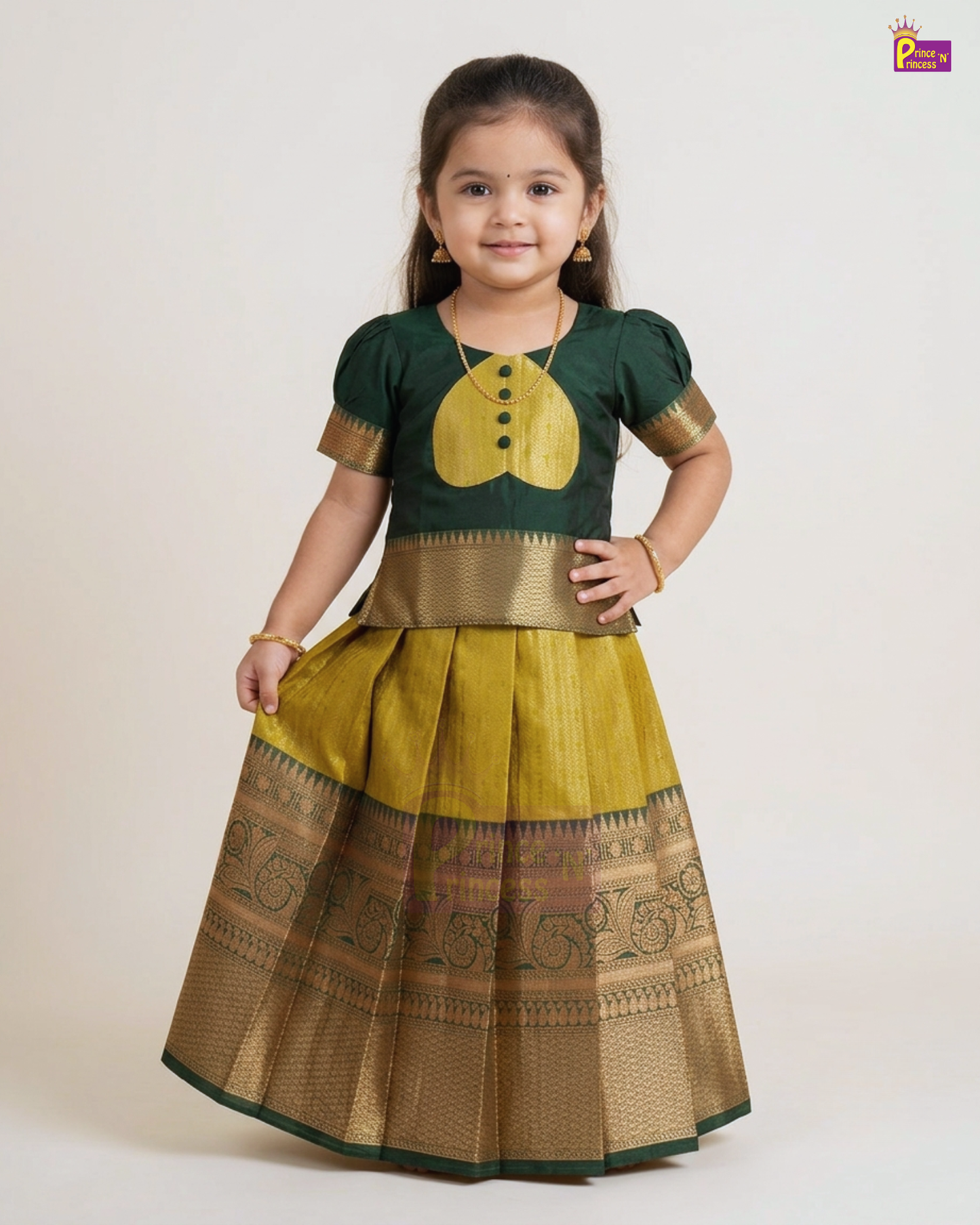 A young girl modeling a traditional Indian ethnic wear set consisting of a dark green short-sleeve blouse with a yellow chest motif and gold trim, paired with a pleated yellow skirt featuring a wide gold zari border.