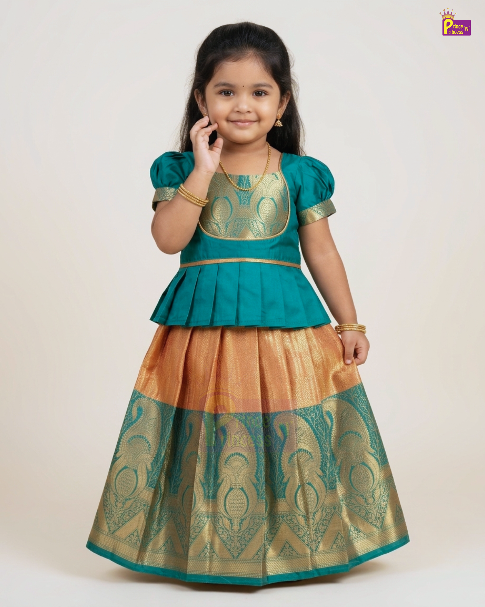 A young girl modeling a traditional Indian lehenga choli dress. The outfit features a teal green peplum-style top with gold embroidery, paired with a flared skirt that transitions from a peach-orange color at the waist to a teal and gold heavily patterned zari border.