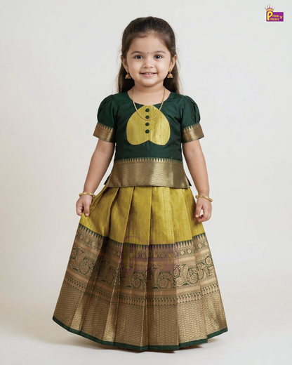 A young girl modeling a traditional Indian ethnic dress consisting of a dark green kurta with a gold yoke and a pleated gold skirt with a wide gold zari border, set against a plain light gray background.