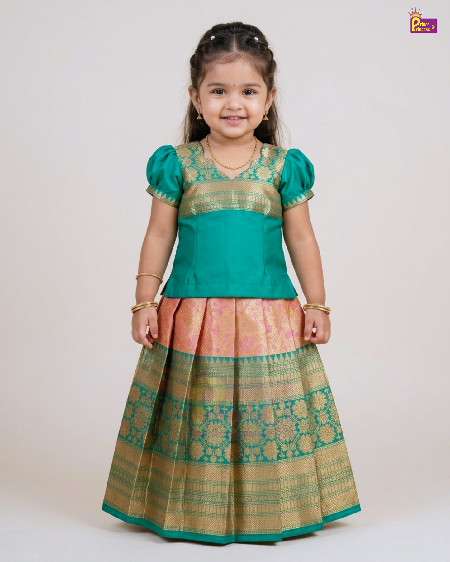 A young girl modeling a traditional Indian ethnic outfit consisting of a teal green top with gold zari embroidery and a matching pleated skirt in pink and green with gold patterns, set against a plain light gray background.