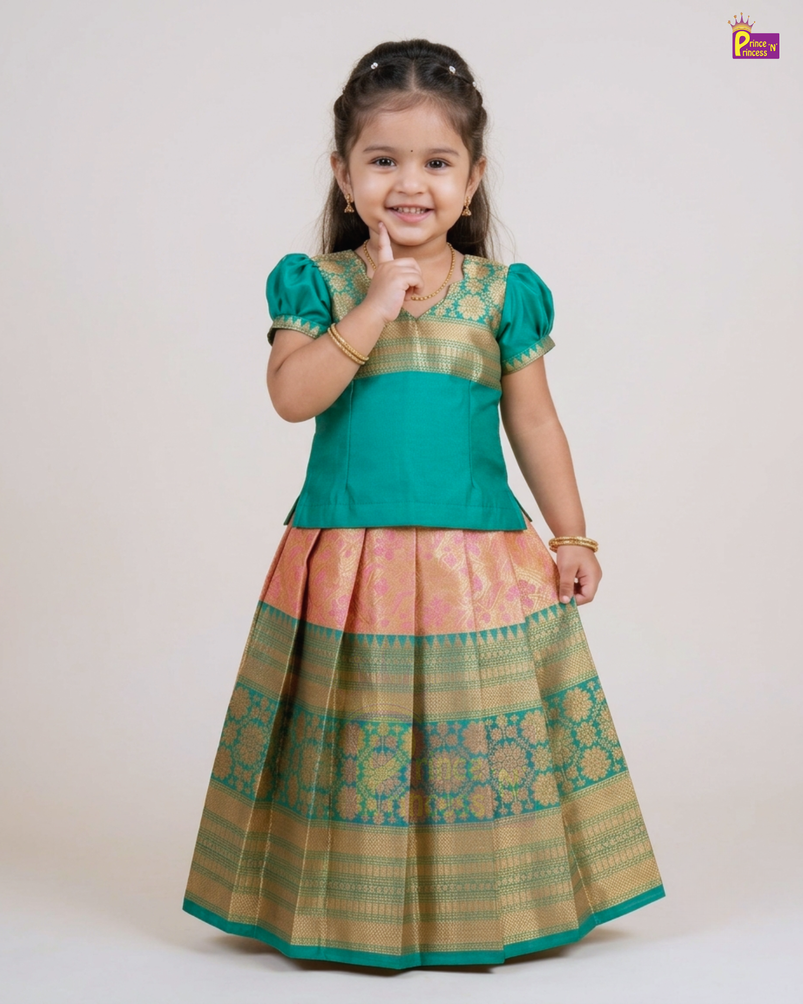 A young girl modeling a traditional Indian ethnic dress with a teal green top and a pleated skirt featuring gold and pink zari patterns, set against a plain light beige background.