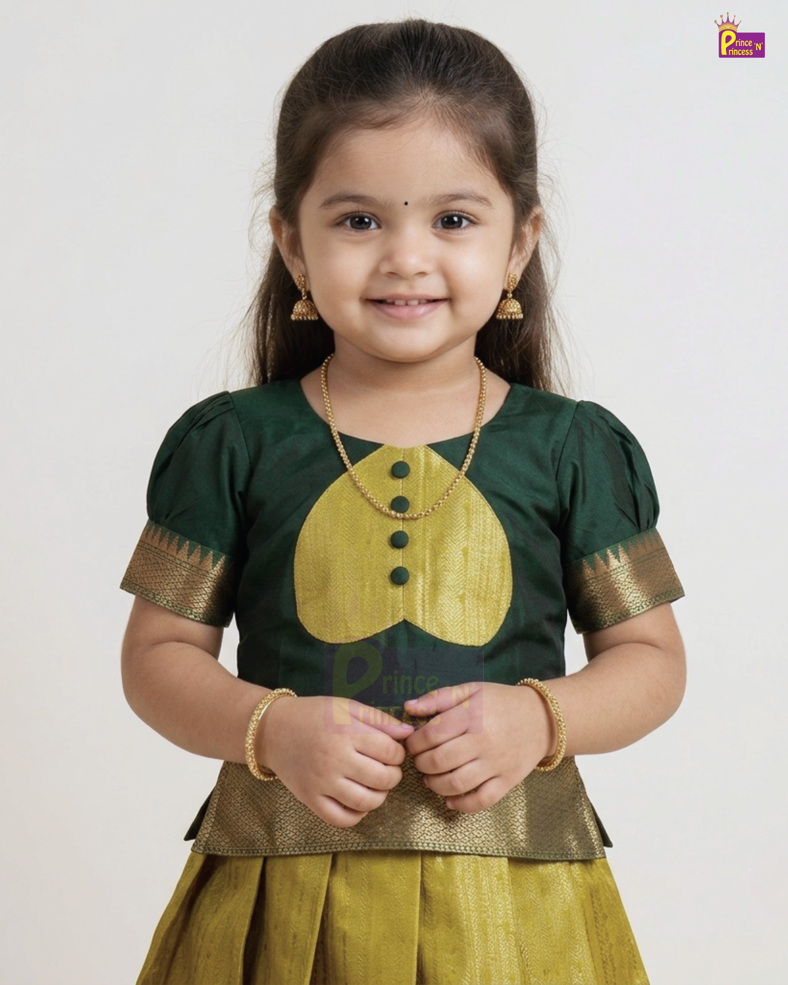 A young girl modeling a traditional Indian outfit consisting of a dark green blouse with a yellow embroidered chest motif and gold-patterned sleeve borders, paired with a matching yellow skirt, against a plain light gray background.