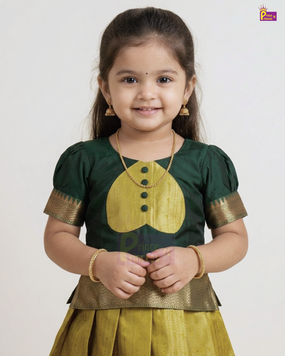 A young girl modeling a traditional Indian outfit consisting of a dark green blouse with a yellow embroidered chest motif and gold-patterned sleeve borders, paired with a matching yellow skirt, against a plain light gray background.