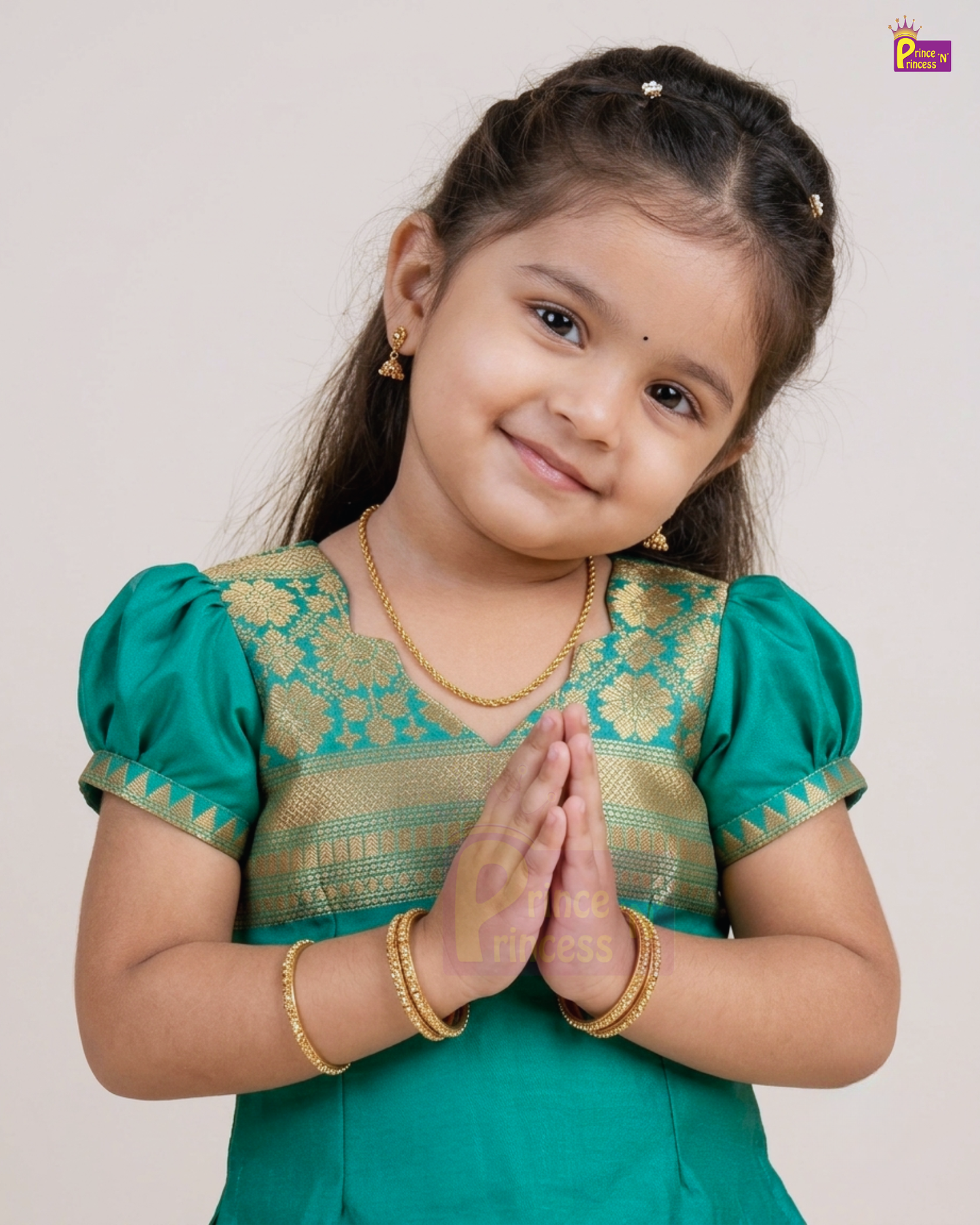 A young girl with dark hair styled in braids, wearing a traditional green and gold embroidered Indian outfit with gold jewelry, posing with her hands together against a plain light background.