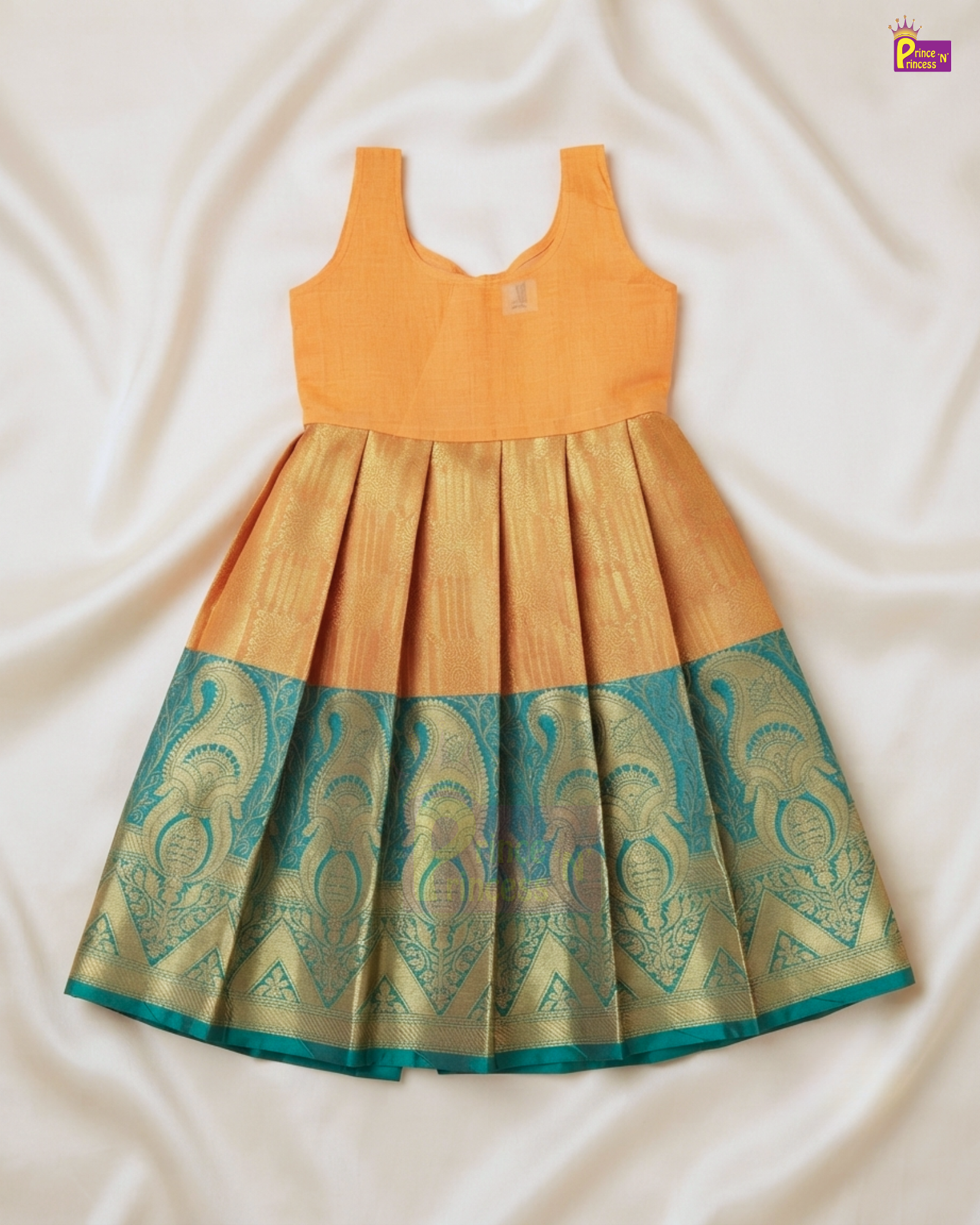 A sleeveless children's dress laid flat on white satin fabric. The dress has a solid mustard yellow-orange bodice and a pleated skirt with a teal and gold woven patterned border.