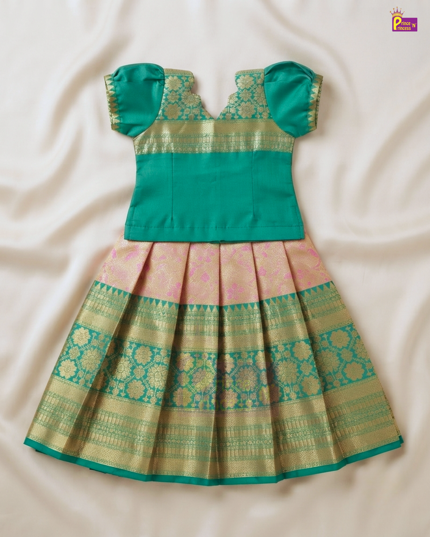 A traditional Indian toddler girl's dress featuring a solid teal green top with puffed sleeves and a pleated skirt in pink and gold zari woven fabric, laid flat on a cream satin background.