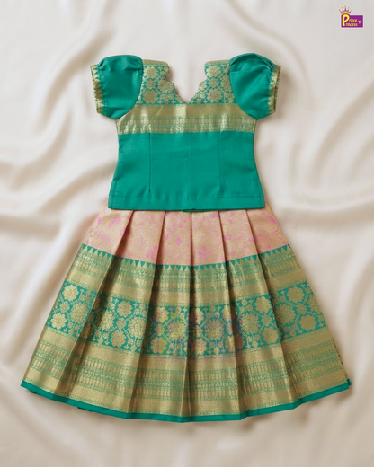 A traditional Indian toddler girl's dress featuring a solid teal green top with puffed sleeves and a pleated skirt in pink and gold zari woven fabric, laid flat on a cream satin background.