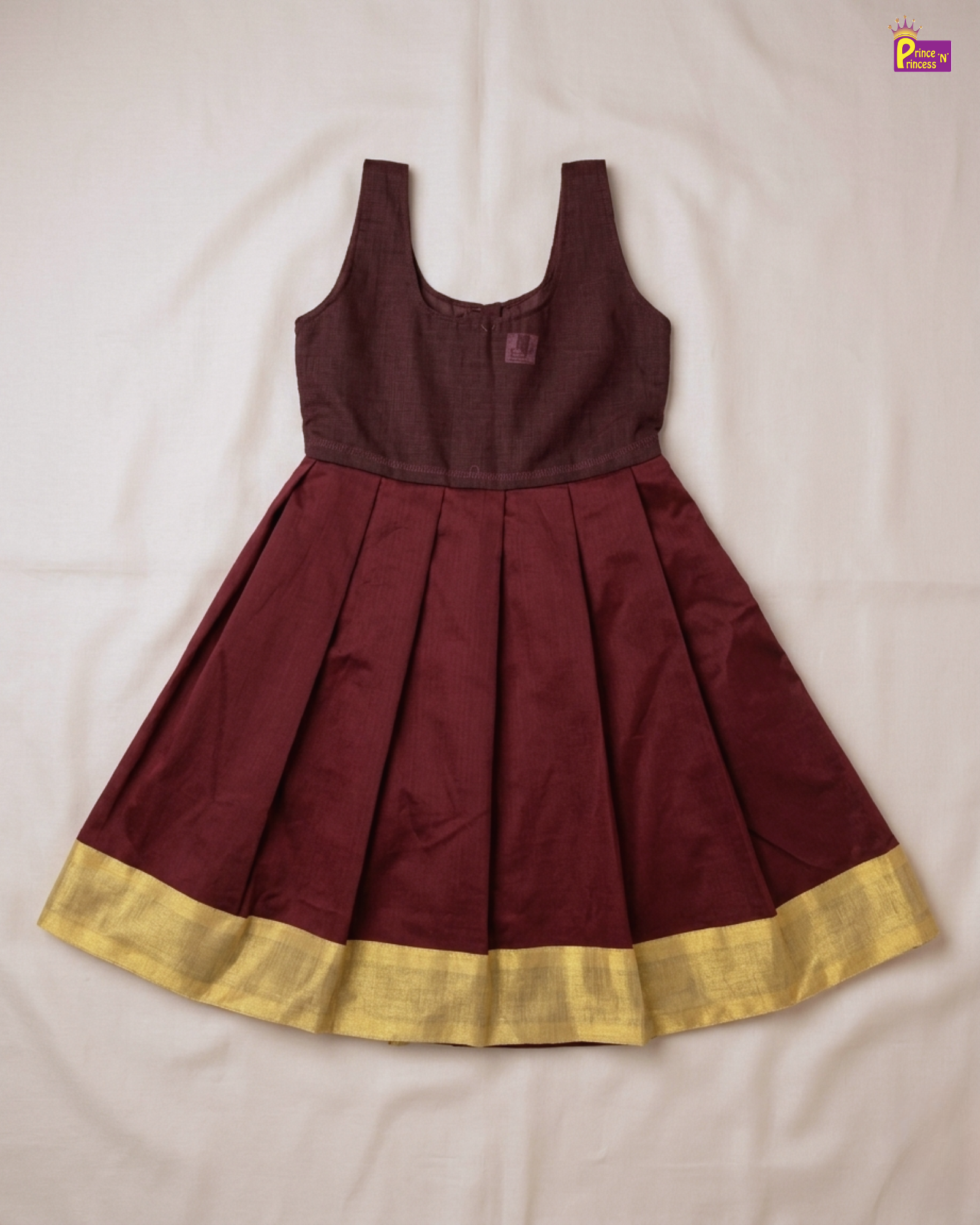A sleeveless traditional-style dress with a dark maroon bodice and a pleated skirt featuring a wide gold border, laid flat on a white fabric background.