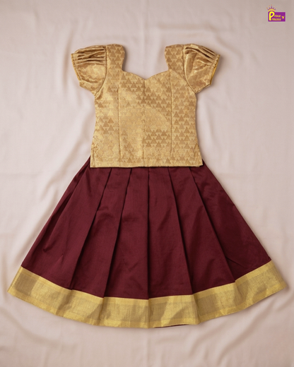 A traditional Indian girl's dress set featuring a gold metallic patterned top with pleated ruffled sleeves and a matching pleated maroon skirt with a gold border, laid flat on a light fabric background.