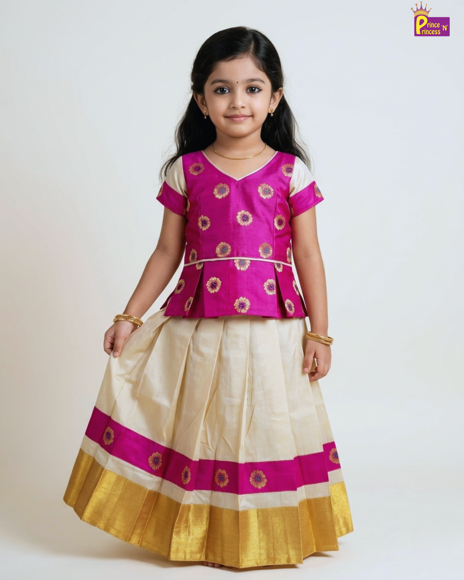 A young girl modeling a traditional Indian outfit consisting of a bright magenta pink peplum top with gold and white floral embroidery, paired with a matching cream-colored skirt that features a wide magenta band and a gold border.