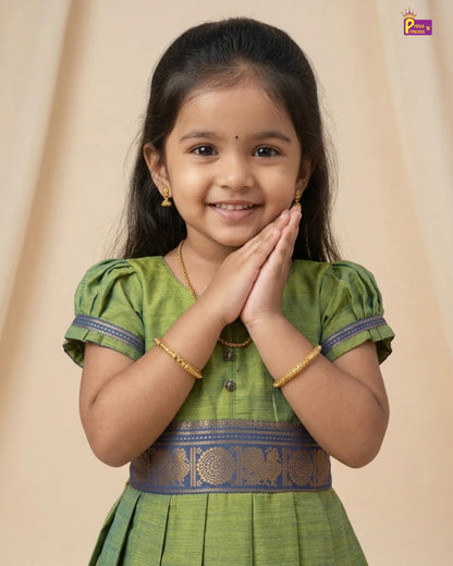 A young girl with dark hair wearing a green traditional Indian dress with gold patterns and blue trim, posing with her hands together in a 'namaste' gesture against a beige background.
