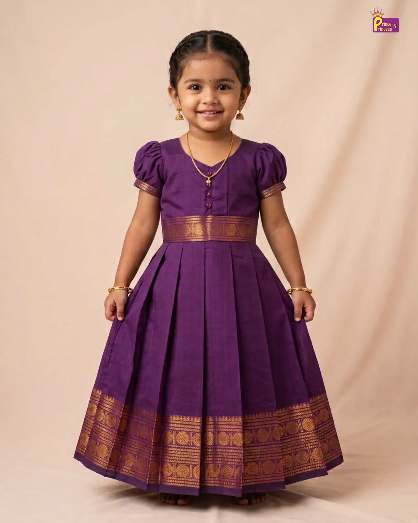 A young girl modeling a traditional purple cotton saree dress with gold zari borders and button details, standing against a beige studio backdrop.