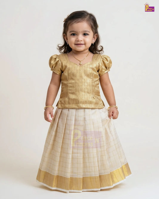 A young girl modeling a traditional Indian outfit consisting of a metallic gold short-sleeve top and a pleated cream and gold skirt, photographed against a plain light background.