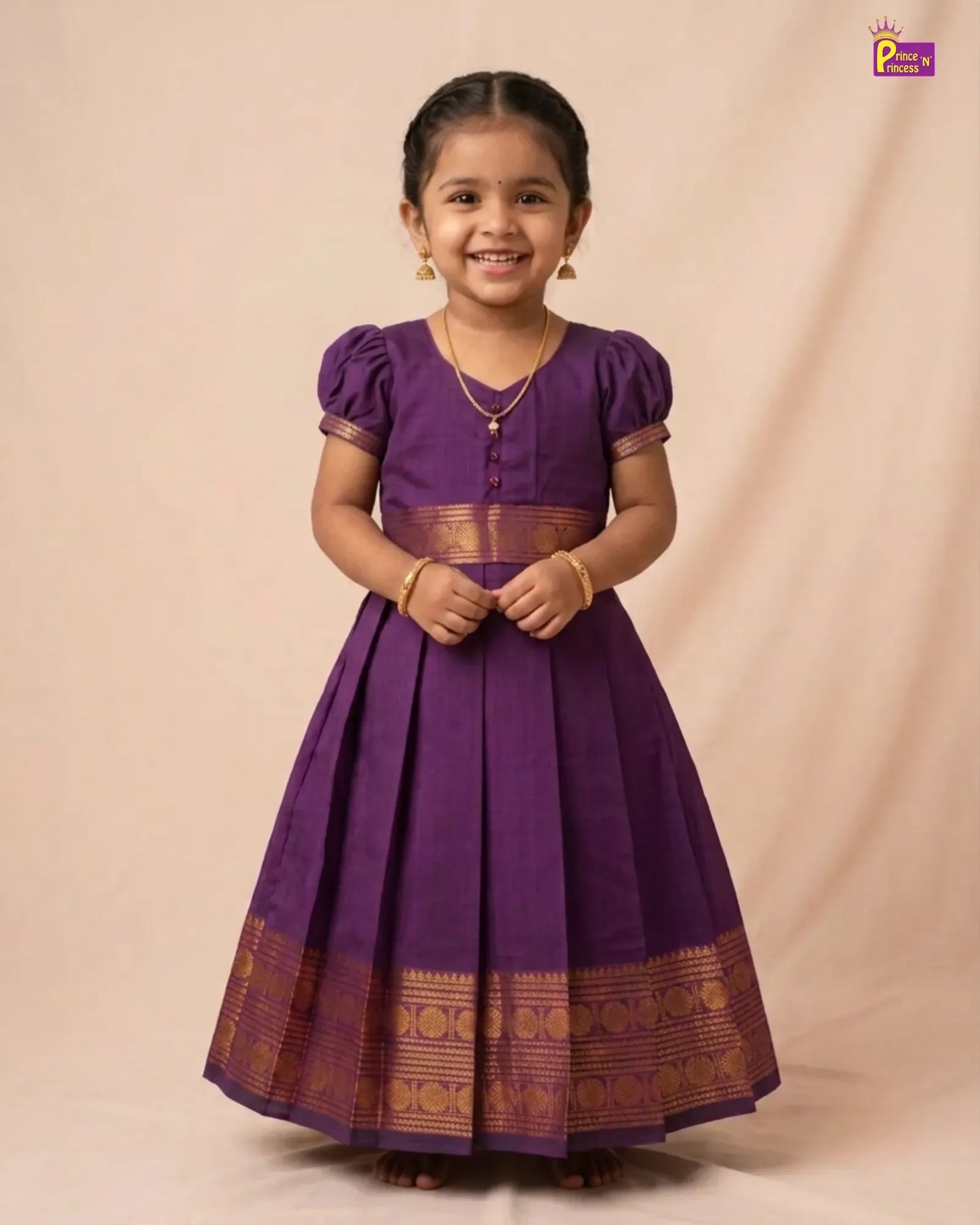 A young girl wearing a purple traditional dress with gold zari borders, standing against a beige studio backdrop.