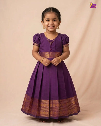 A young girl wearing a purple traditional dress with gold zari borders, standing against a beige studio backdrop.