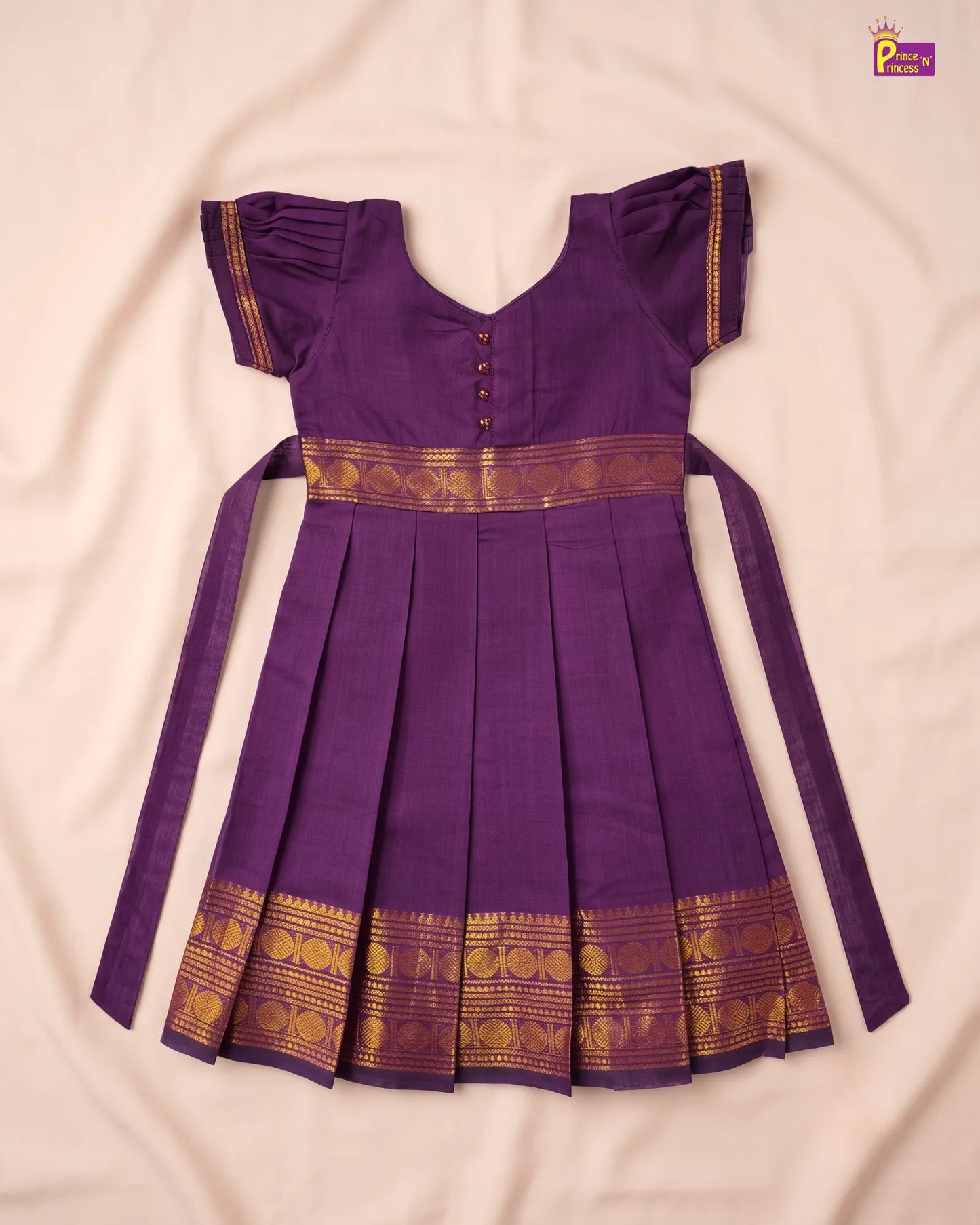 A purple traditional Indian dress with gold zari borders, laid flat on a light beige fabric background. The dress features short flutter sleeves, a V-neckline with decorative buttons, a pleated skirt, and long side ties.