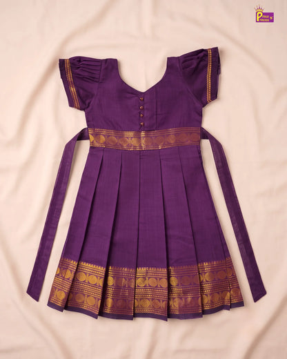 A purple traditional Indian dress with gold zari borders, laid flat on a light beige fabric background. The dress features short flutter sleeves, a V-neckline with decorative buttons, a pleated skirt, and long side ties.