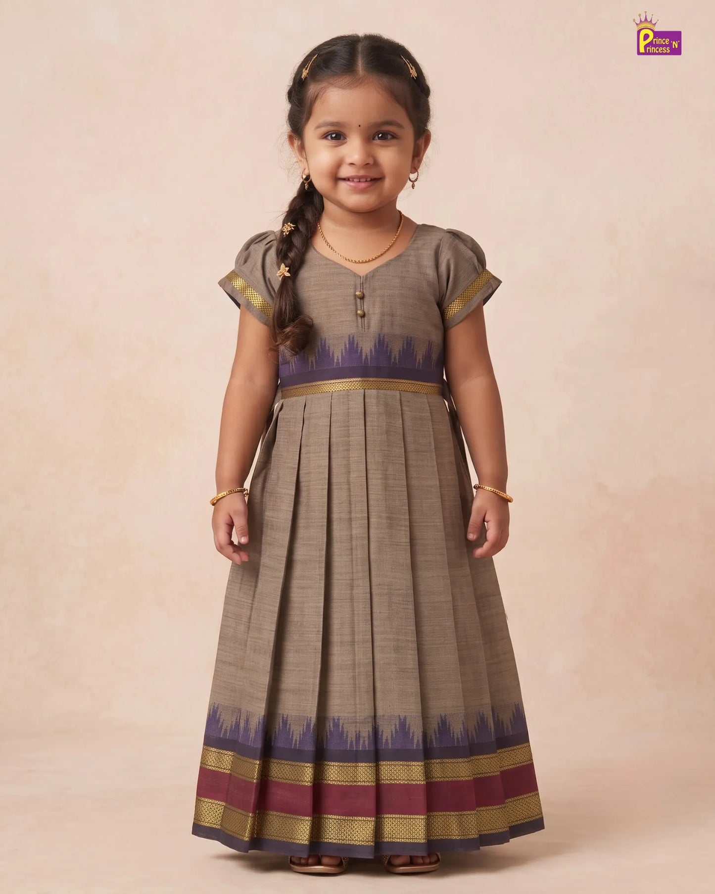 A young girl with a braid stands against a warm beige studio backdrop wearing a long pleated dress. The dress is taupe-brown with a purple and gold patterned hem and waistband.