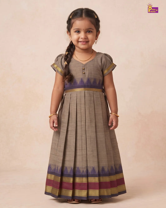 A young girl with a braid stands against a warm beige studio backdrop wearing a long pleated dress. The dress is taupe-brown with a purple and gold patterned hem and waistband.