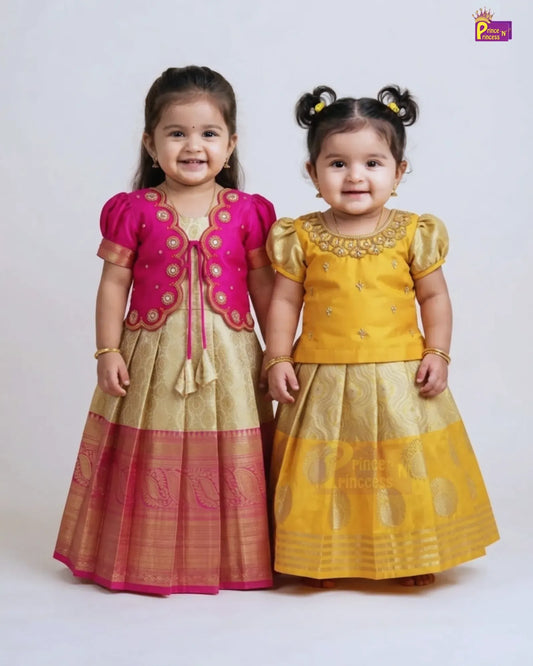 Two young girls modeling traditional Indian ethnic wear, standing side-by-side against a light gray studio background. The girl on the left wears a pink and gold embroidered kurta and skirt set, while the girl on the right wears a yellow and gold embroidered kurta and skirt set.