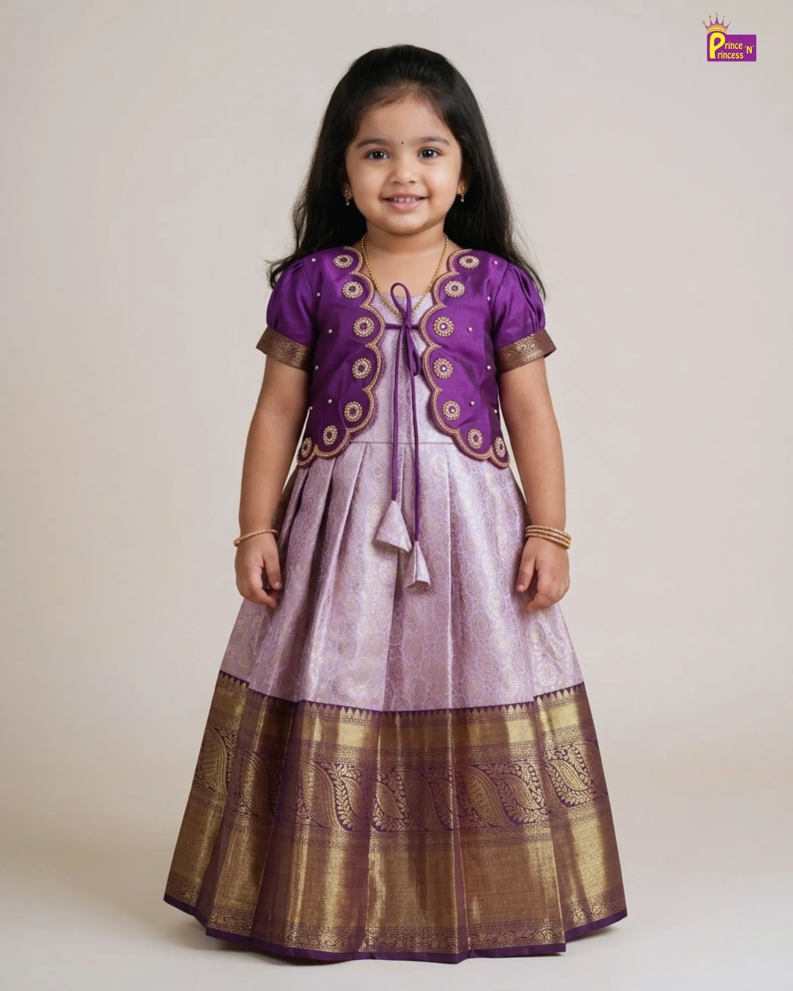 A young girl modeling a traditional Indian ethnic dress with a purple and lavender skirt, decorated with gold zari embroidery and a matching purple embroidered jacket. The image is shot against a plain beige studio background.