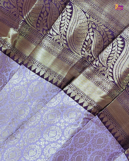 A close-up of a woven fabric, likely a sari, featuring a lavender purple body with metallic gold floral motifs and a wide gold zari border with matching paisley patterns.