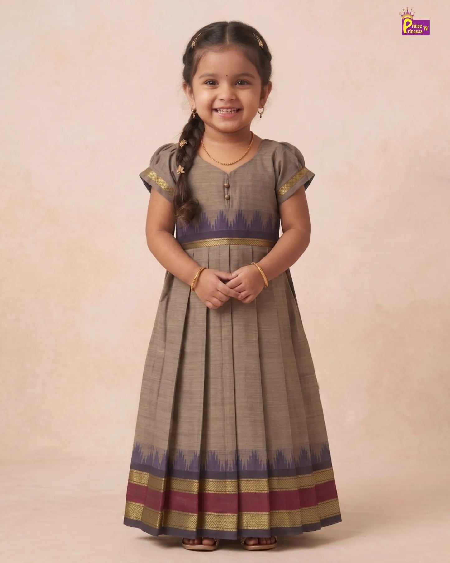 A young girl with braided hair stands wearing a traditional pleated Anarkali-style dress. The dress is primarily taupe brown with a purple and gold patterned waistband and hem, set against a warm beige studio backdrop.