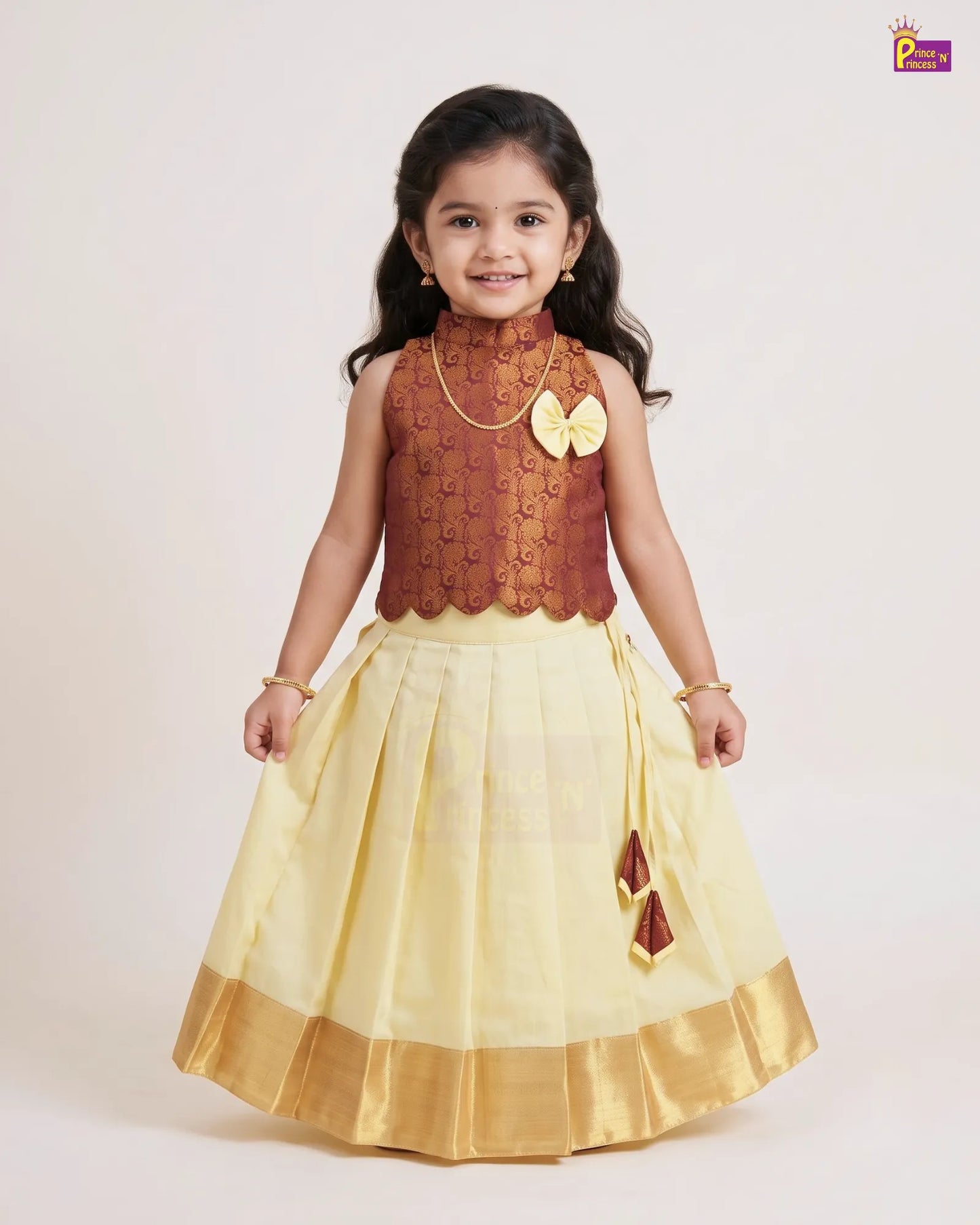 A young girl modeling a traditional Indian outfit consisting of a brown patterned sleeveless top and a pleated light yellow skirt with a gold border, set against a plain light background.