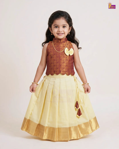 A young girl modeling a traditional Indian outfit consisting of a brown patterned sleeveless top and a pleated light yellow skirt with a gold border, set against a plain light background.