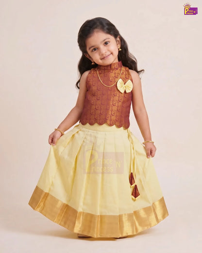 A young girl modeling a traditional Indian outfit consisting of a brown embroidered sleeveless top with a yellow bow accent and a light yellow flared skirt with a gold border, set against a plain beige background.