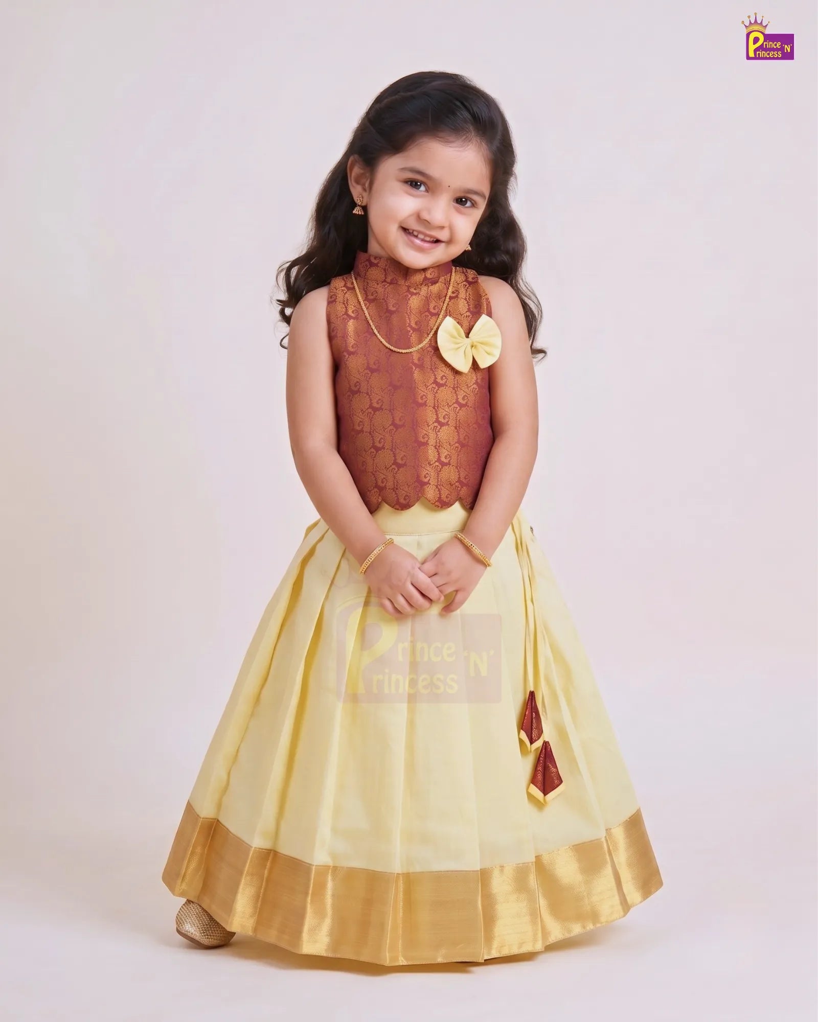 A young girl modeling a traditional Indian ethnic dress with a brown embroidered bodice and a pleated light yellow skirt with a gold border, set against a plain light background.